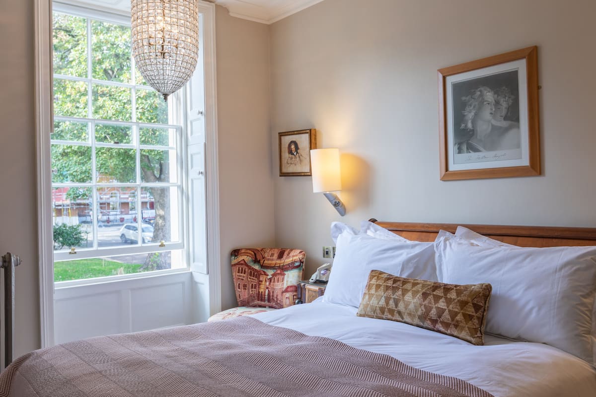 A bedroom with a large window, a chandelier, a bed with white and patterned bedding, a nightstand with a lamp, and a framed picture on the wall.