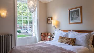 Een slaapkamer met een groot raam, een kroonluchter, een bed met witte en gestreepte beddengoed, een nachtkastje met een lamp, en een ingelijst schilderij aan de muur.