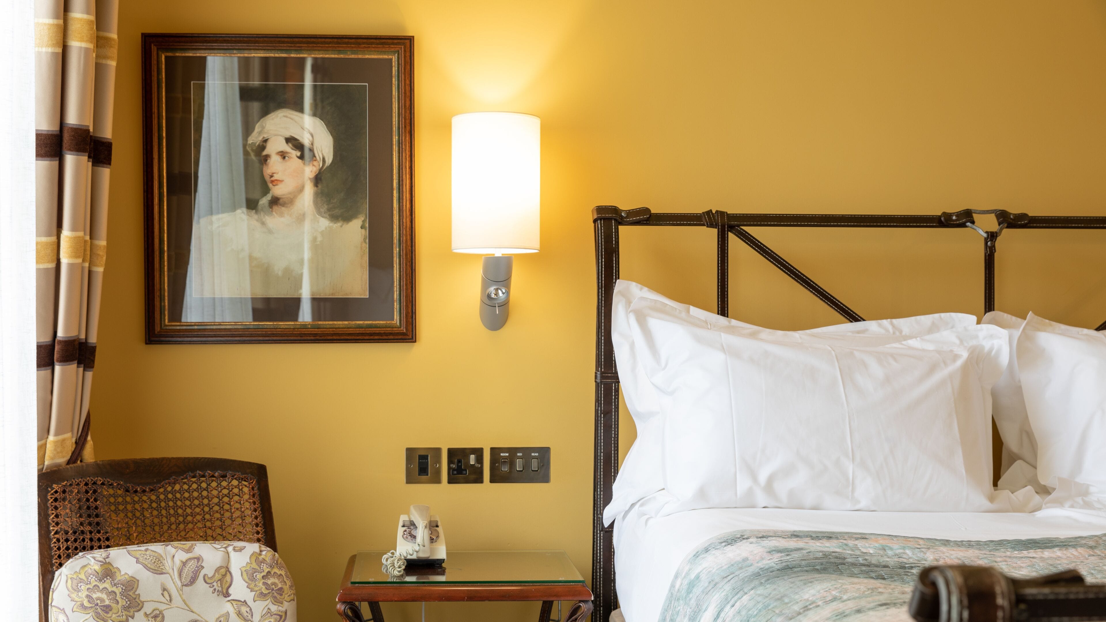 A hotel room with a bed, a bedside table with a telephone, a framed portrait on the wall, and a lamp.