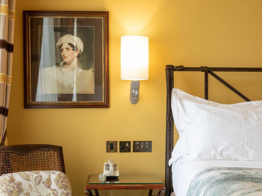 A hotel room with a bed, a bedside table with a telephone, a framed portrait on the wall, and a lamp.