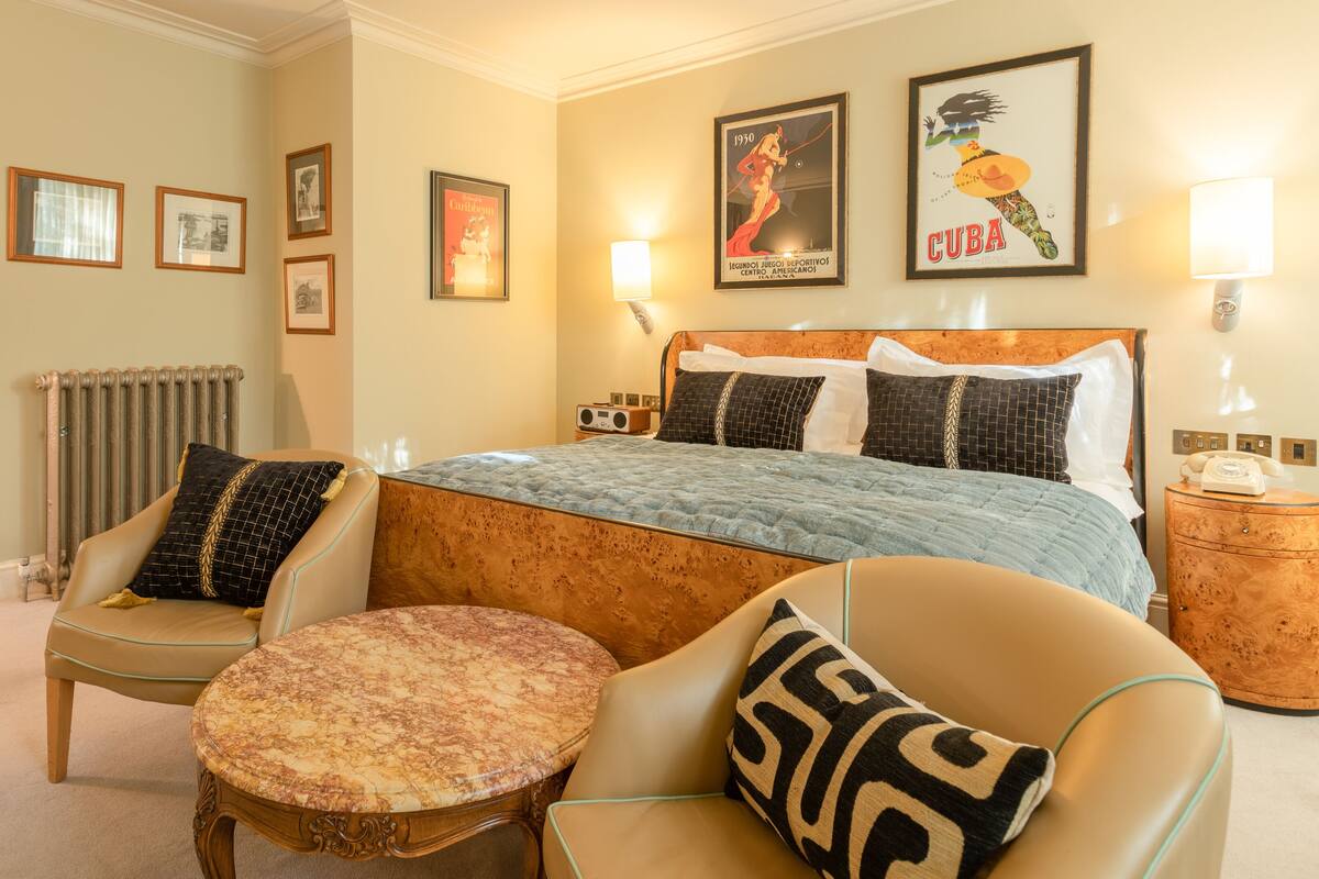 A hotel room with a large bed, two chairs, a small table, and a radiator. There are framed pictures on the wall and a lamp on the nightstand.