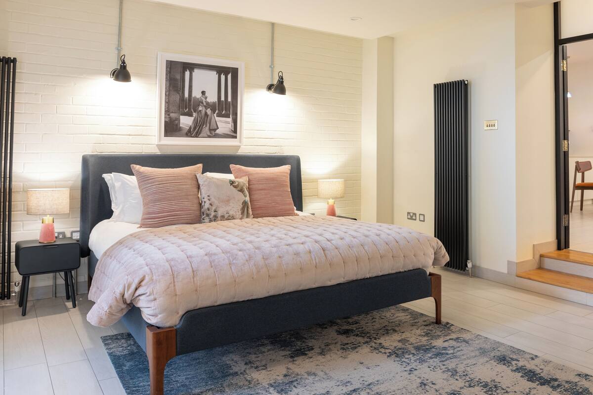 A modern bedroom with a large bed, bedside lamps, a framed picture on the wall, and a view into the dining area.