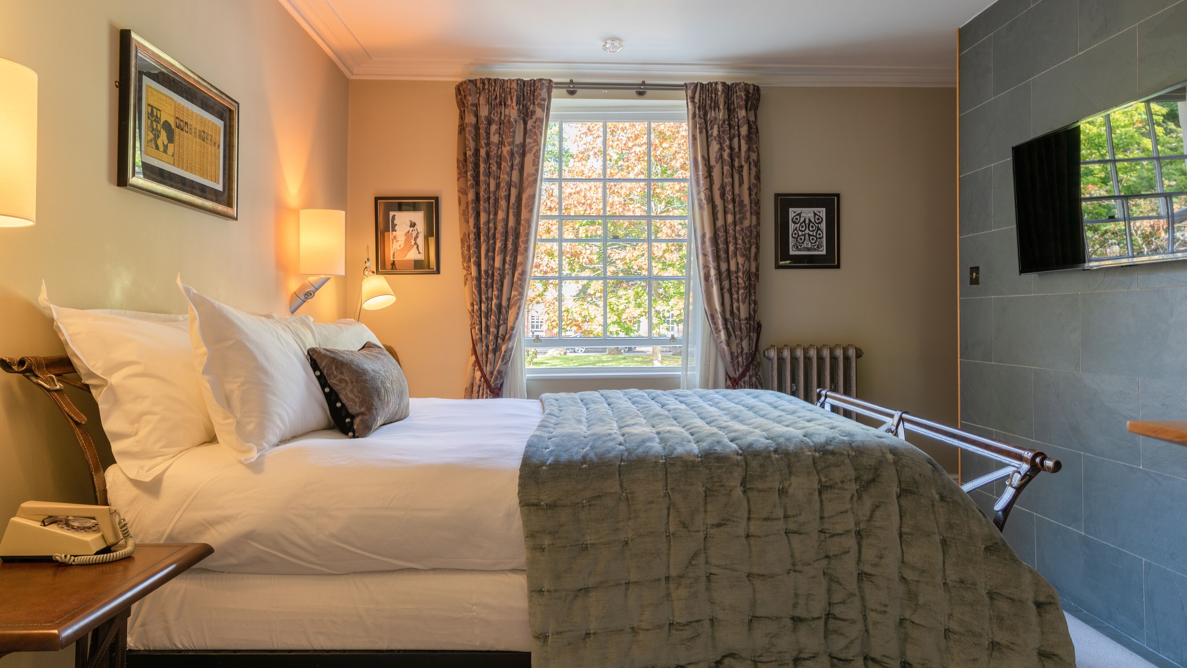 A hotel room with a bed, a bedside telephone, a window with curtains, a radiator, and framed pictures on the walls.