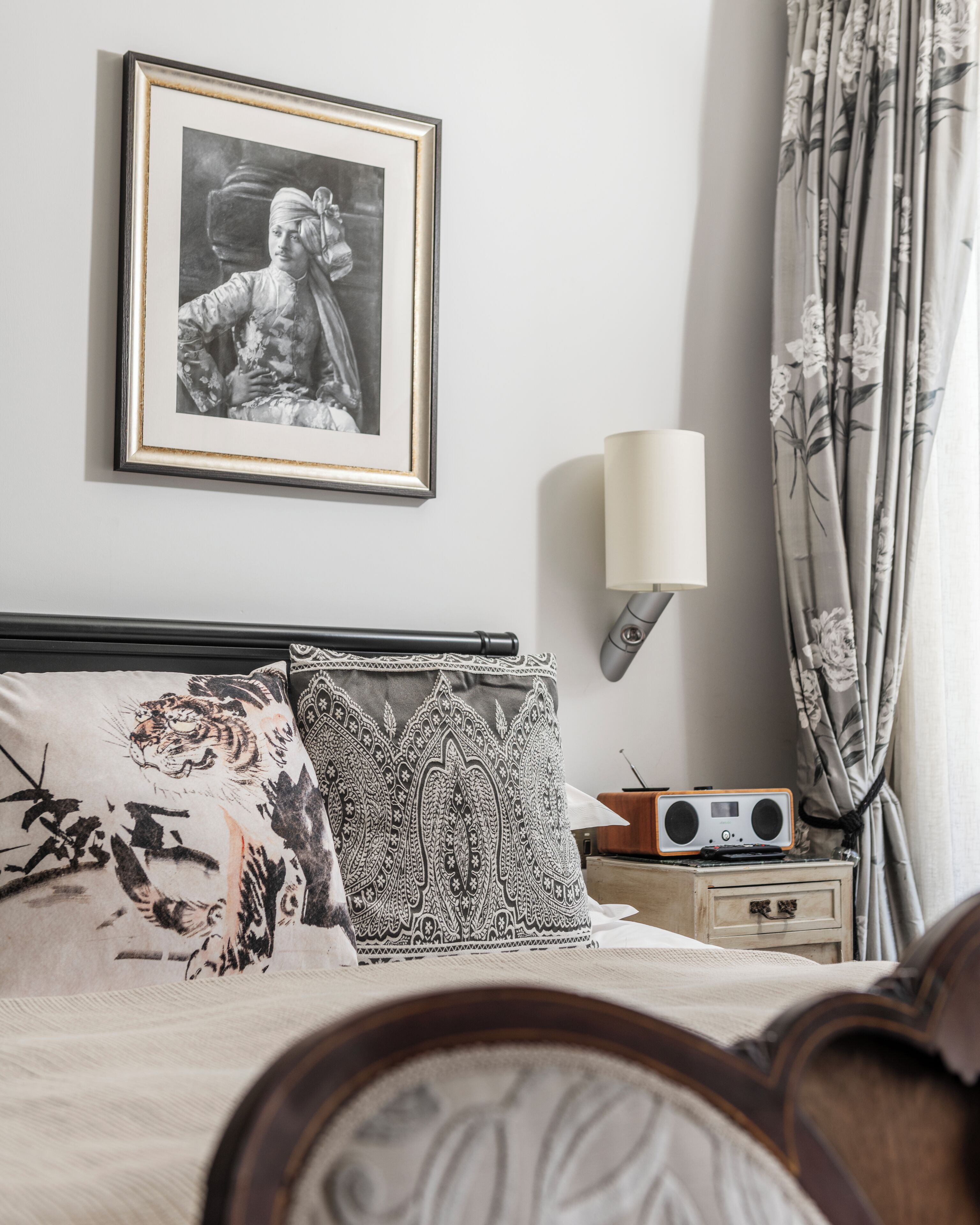 Quarto com cama, almofadas, quadro emoldurado, abajur, rádio e cortinas.