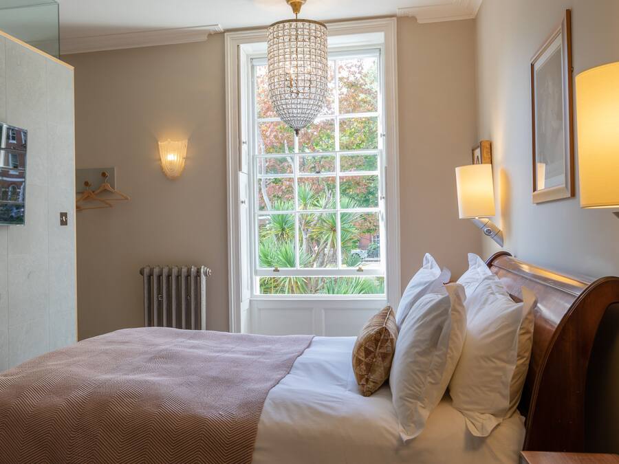 Quarto com cama, radiador, janela com vista para árvores, lustre e televisão fixada na parede.
