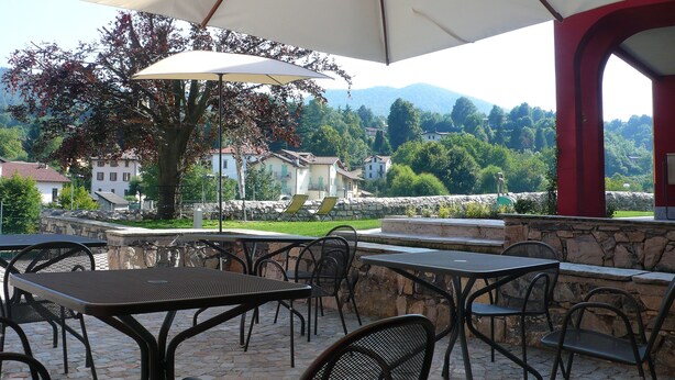 Un patio al aire libre con suelo de piedra, mesas y sillas de metal, y una sombrilla grande que proporciona sombra.