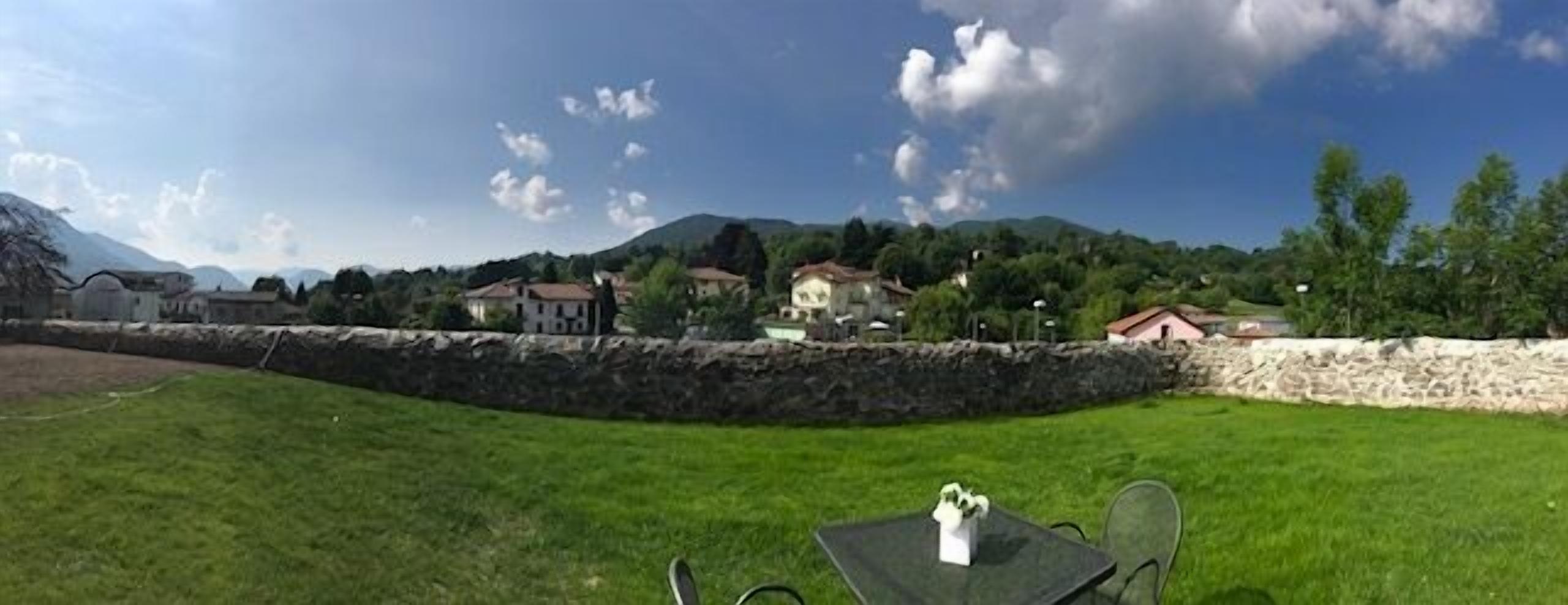 A grassy outdoor area with a stone wall, a table with chairs, and buildings in the background.