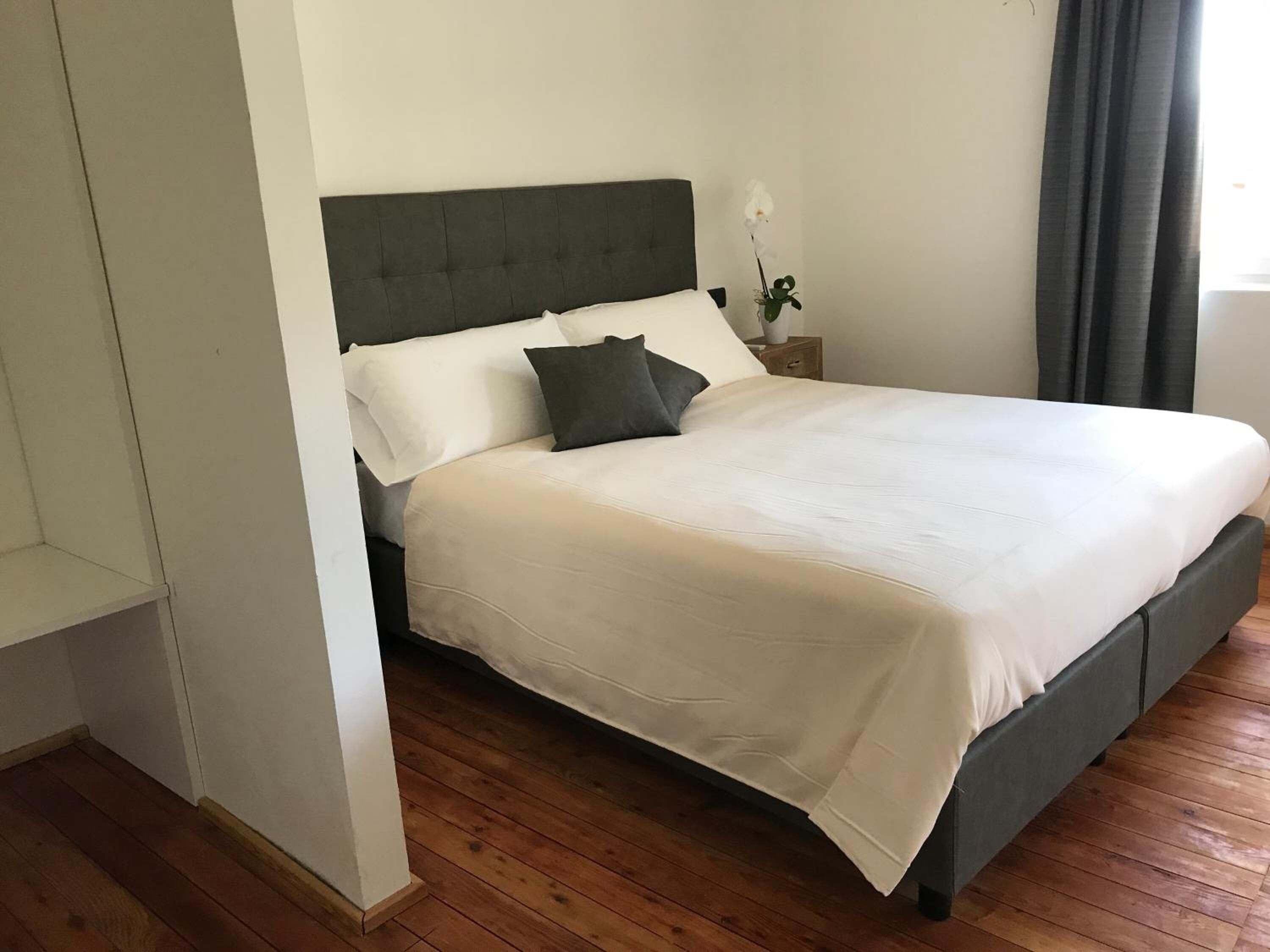 A neatly made bed with a grey headboard and white bedding, a wooden floor, and a small bedside table with a vase of flowers.