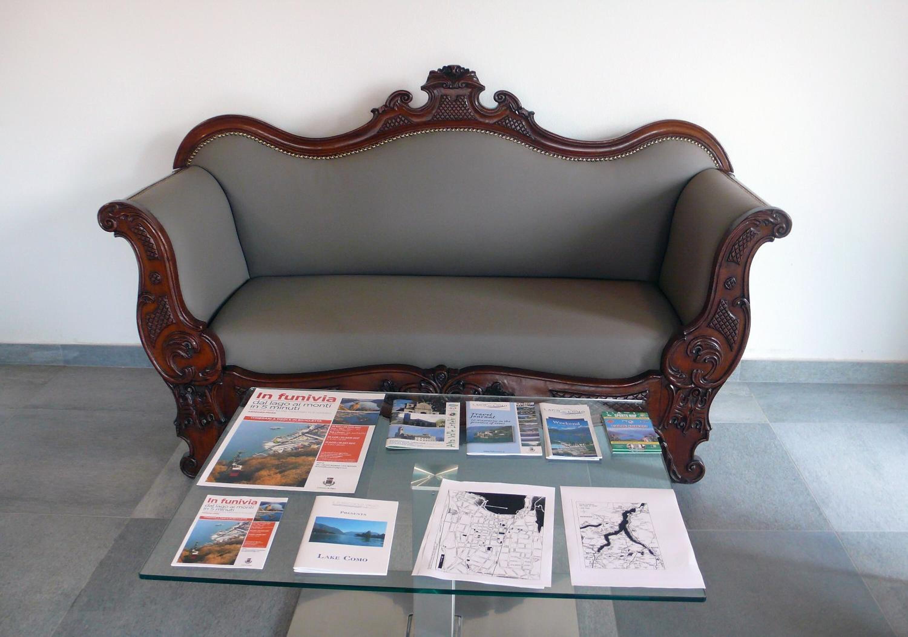 Um sofá antigo com esculturas em madeira elaboradas, uma mesa de vidro com folhetos e mapas, e uma parede branca simples ao fundo.