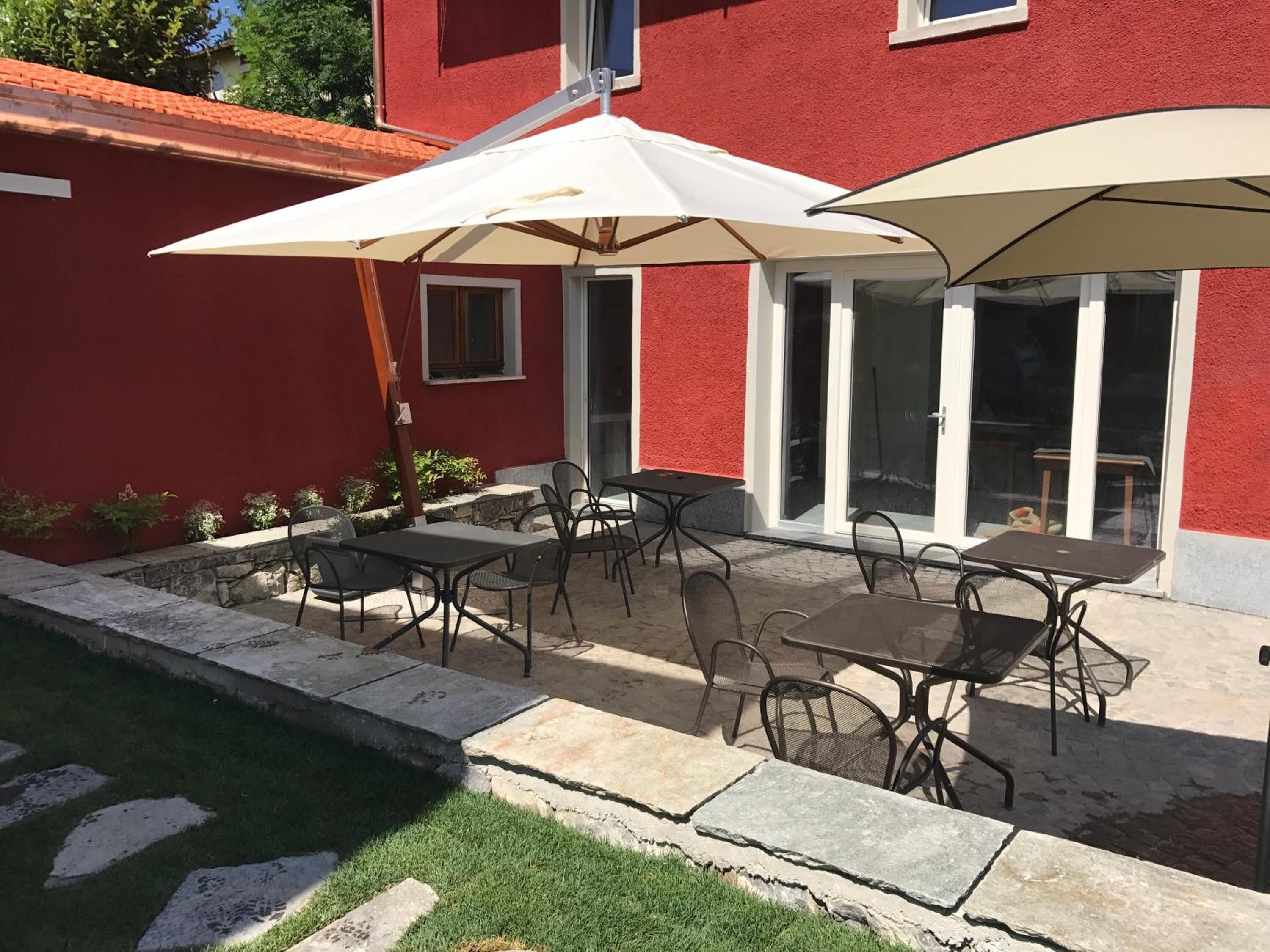 Una terrazza esterna con tavoli e sedie, un ombrellone bianco e un edificio rosso sullo sfondo.