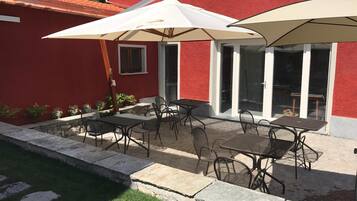 Una terrazza esterna con tavoli e sedie, un ombrellone bianco e un edificio rosso sullo sfondo.