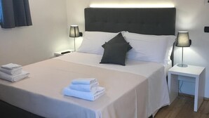 Una cama bien hecha con ropa de cama blanca y gris, dos lámparas de noche y un piso de madera.