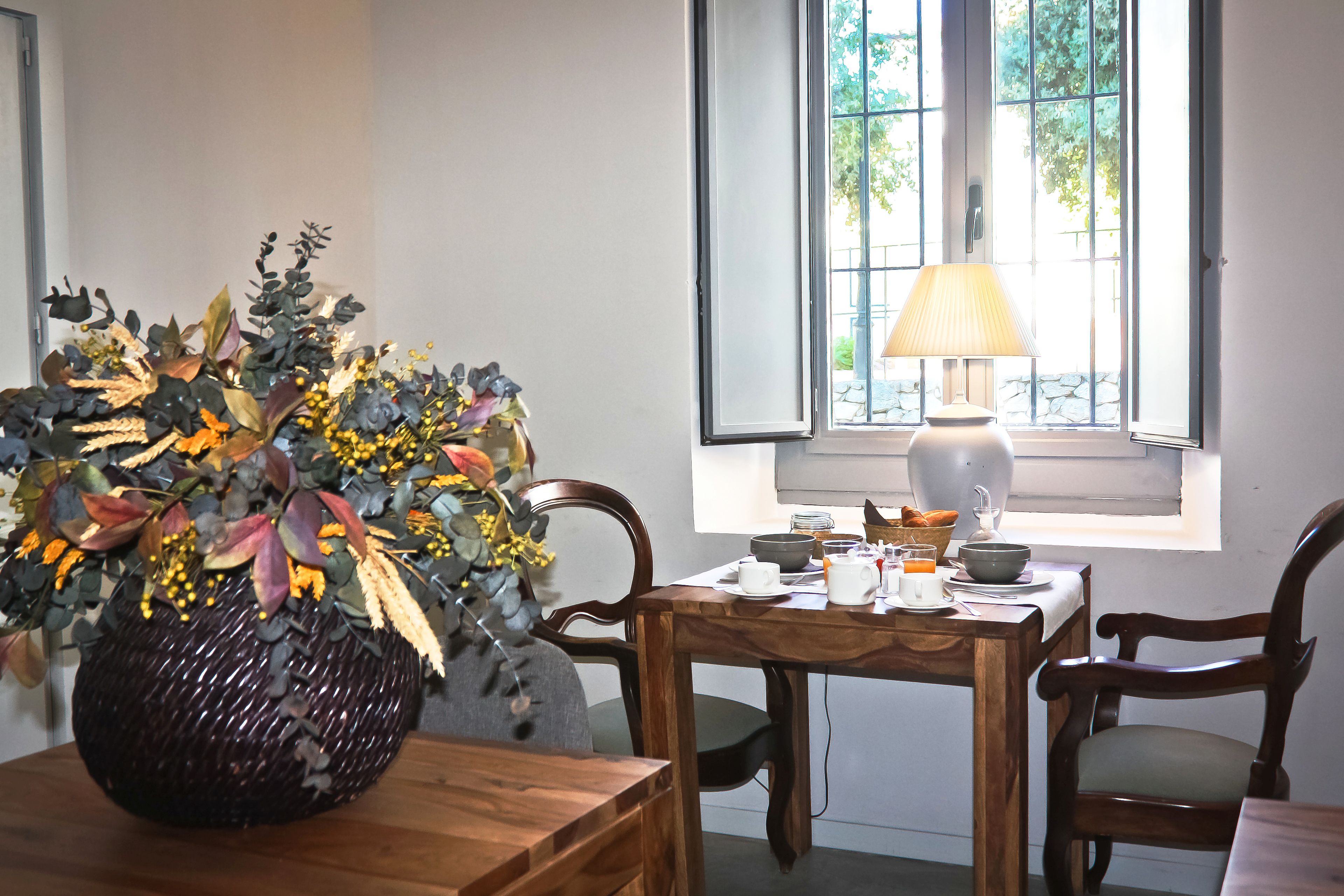 Un coin repas avec une table en bois, des chaises, un vase rempli de feuilles d’automne, une lampe et une fenêtre donnant sur un paysage verdoyant.