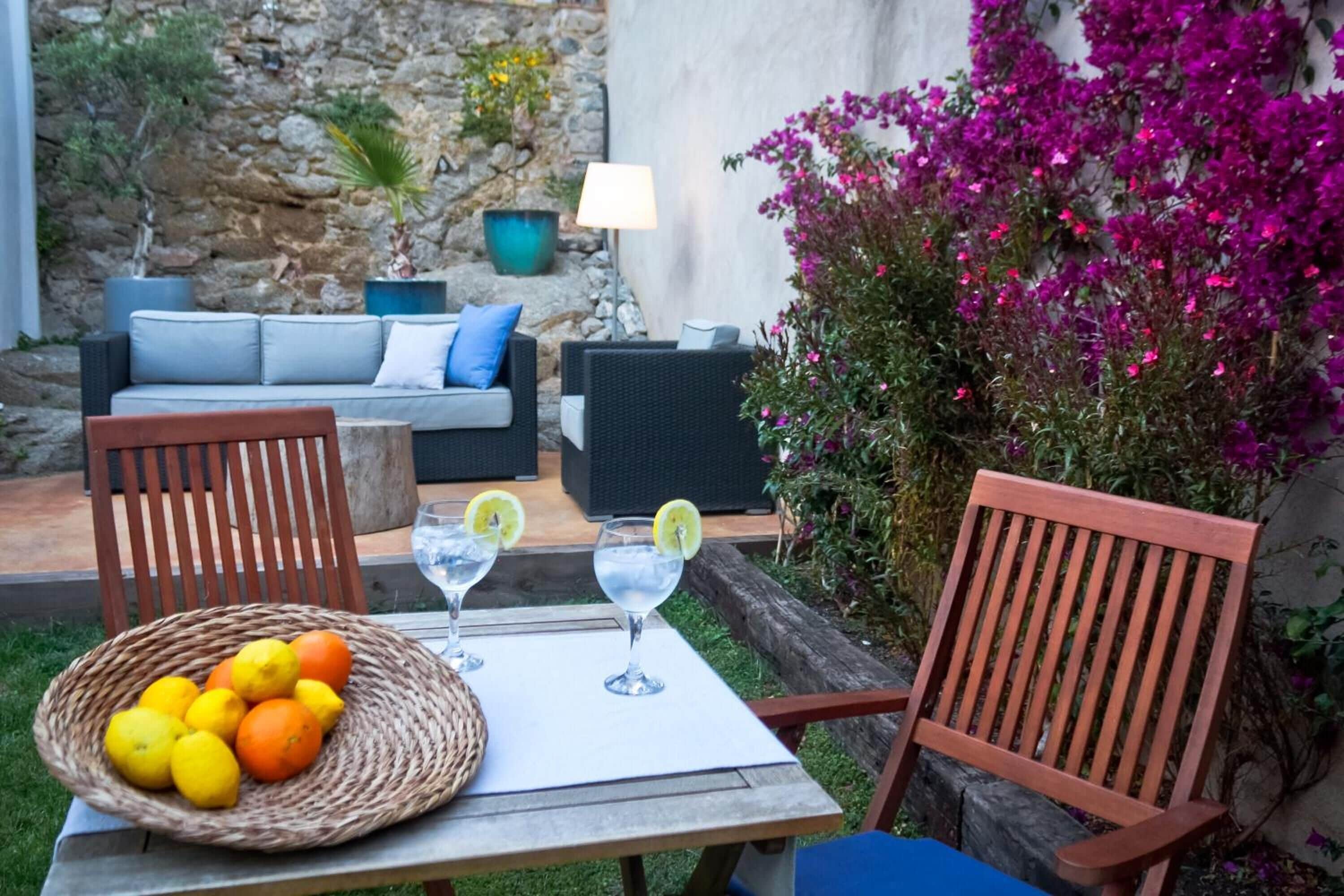 Um pátio com cadeiras de vime, uma mesa com bebidas e frutas, e uma parede de pedra com plantas.