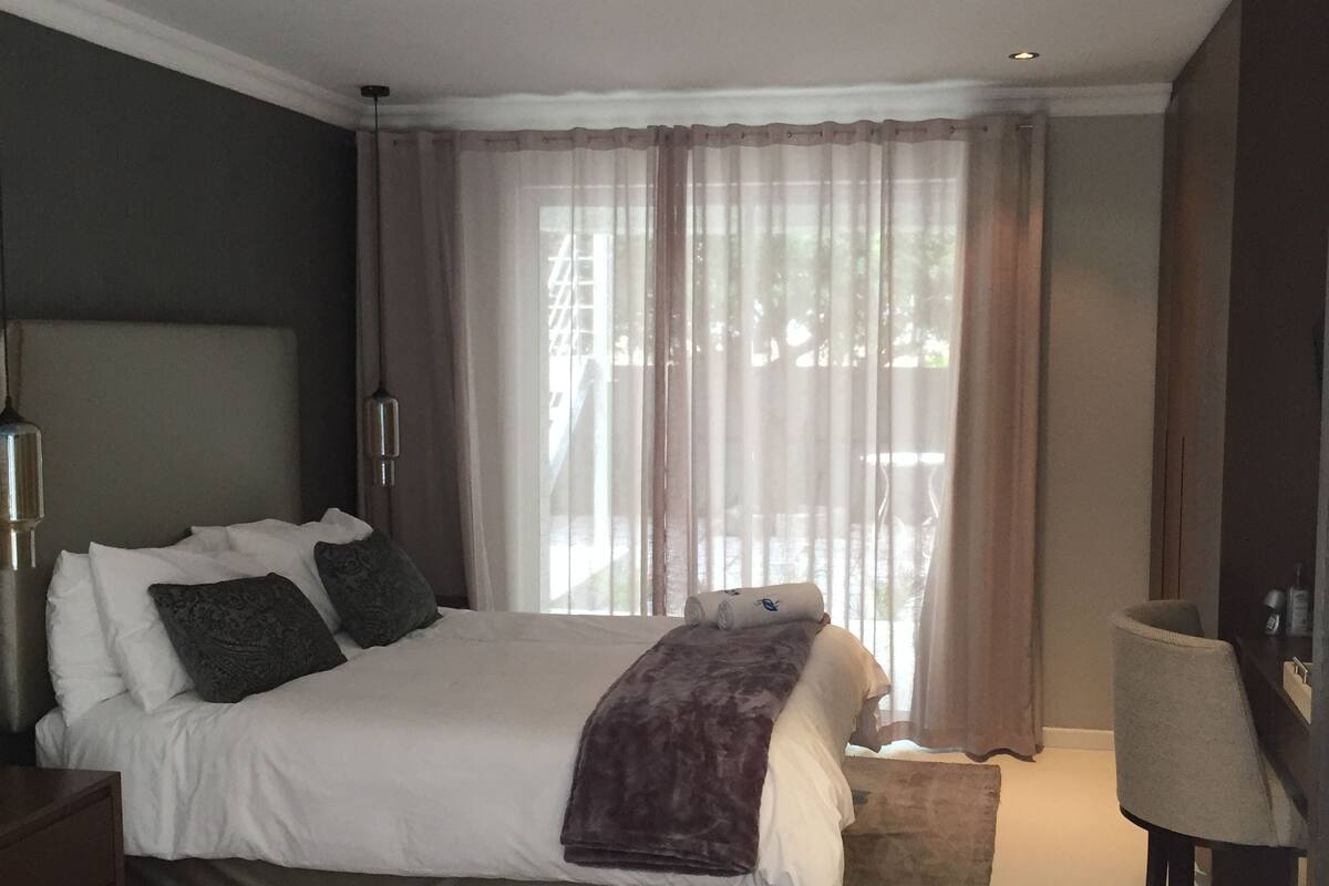A modern bedroom with a large bed, a bedside lamp, and a view of the outdoors through sheer curtains.