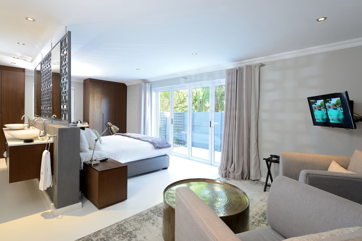A modern hotel room with a large bed, a sitting area with a sofa and a coffee table, a TV mounted on the wall, and a balcony with curtains.