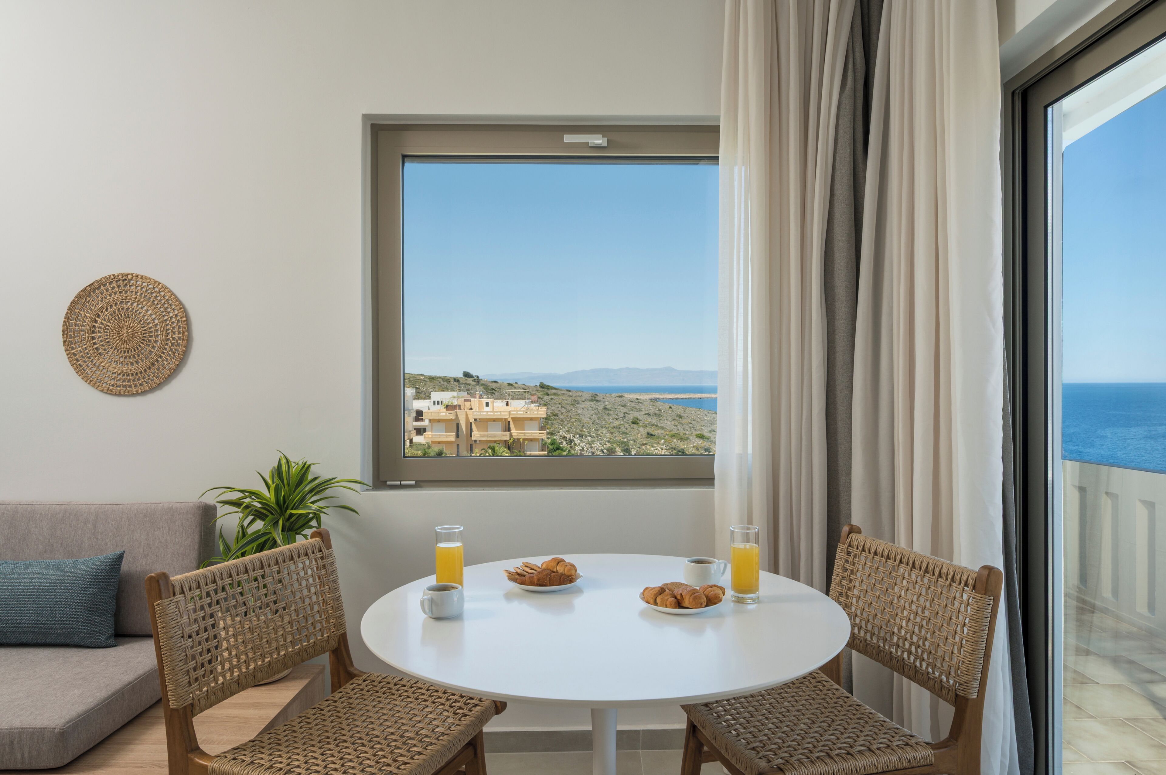 A room with a round table, two wicker chairs, a sofa, and a view of the sea through a large window.