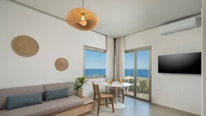 A modern living room with a sofa, dining table, and chairs, a TV mounted on the wall, and a balcony with a view. - Esplanade Apartments (Chania)