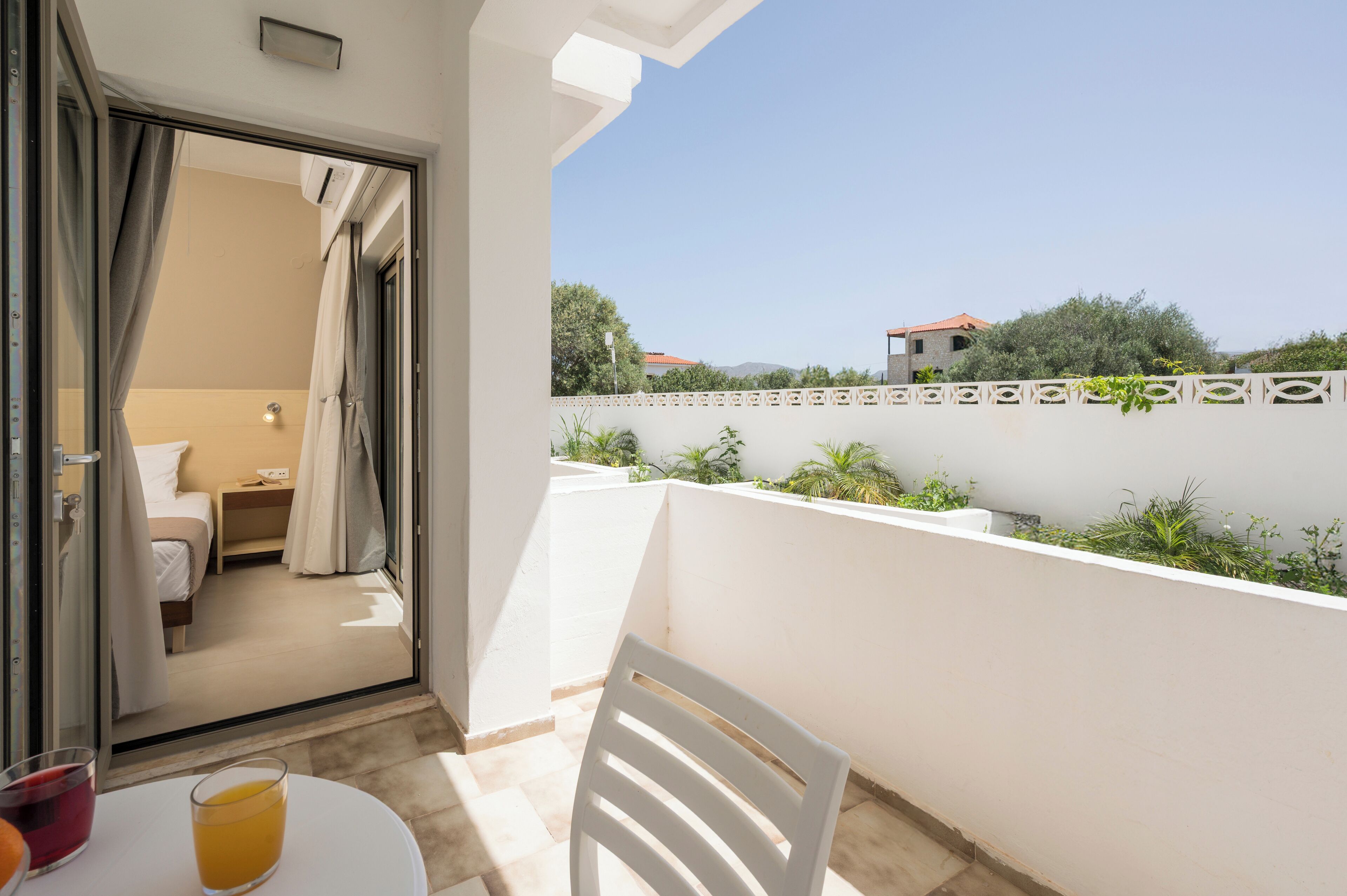 A balcony with a white chair, a table with a glass of orange juice, and a view of a bedroom with a bed and nightstand.