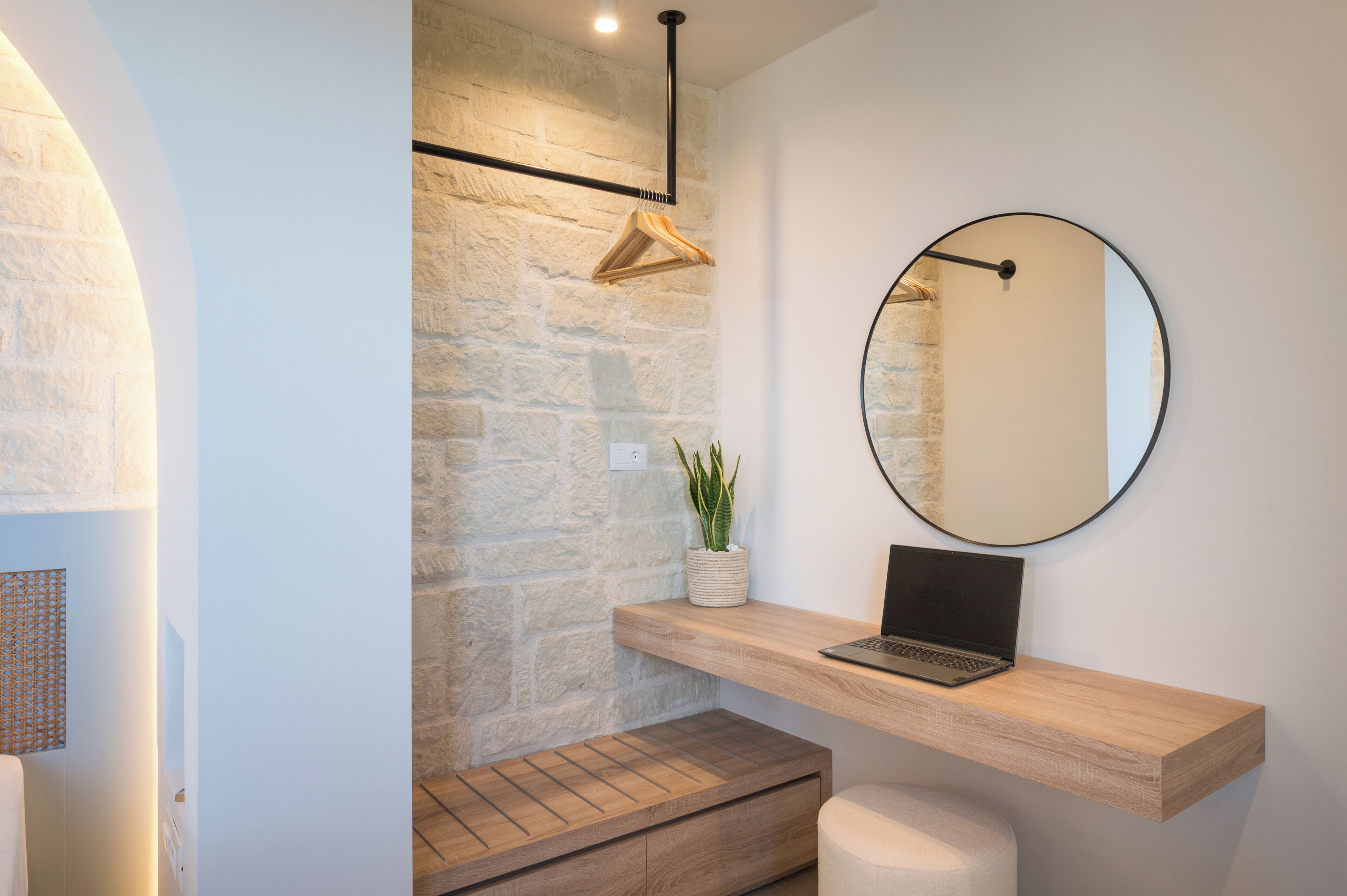 A modern bathroom with a stone wall, a wooden vanity, a round mirror, and a laptop on the vanity.