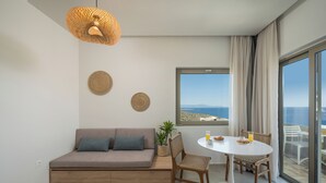 A modern living room with a sofa, dining table, chairs, a hanging light fixture, and a large window with a view. - Esplanade Apartments (Chania)