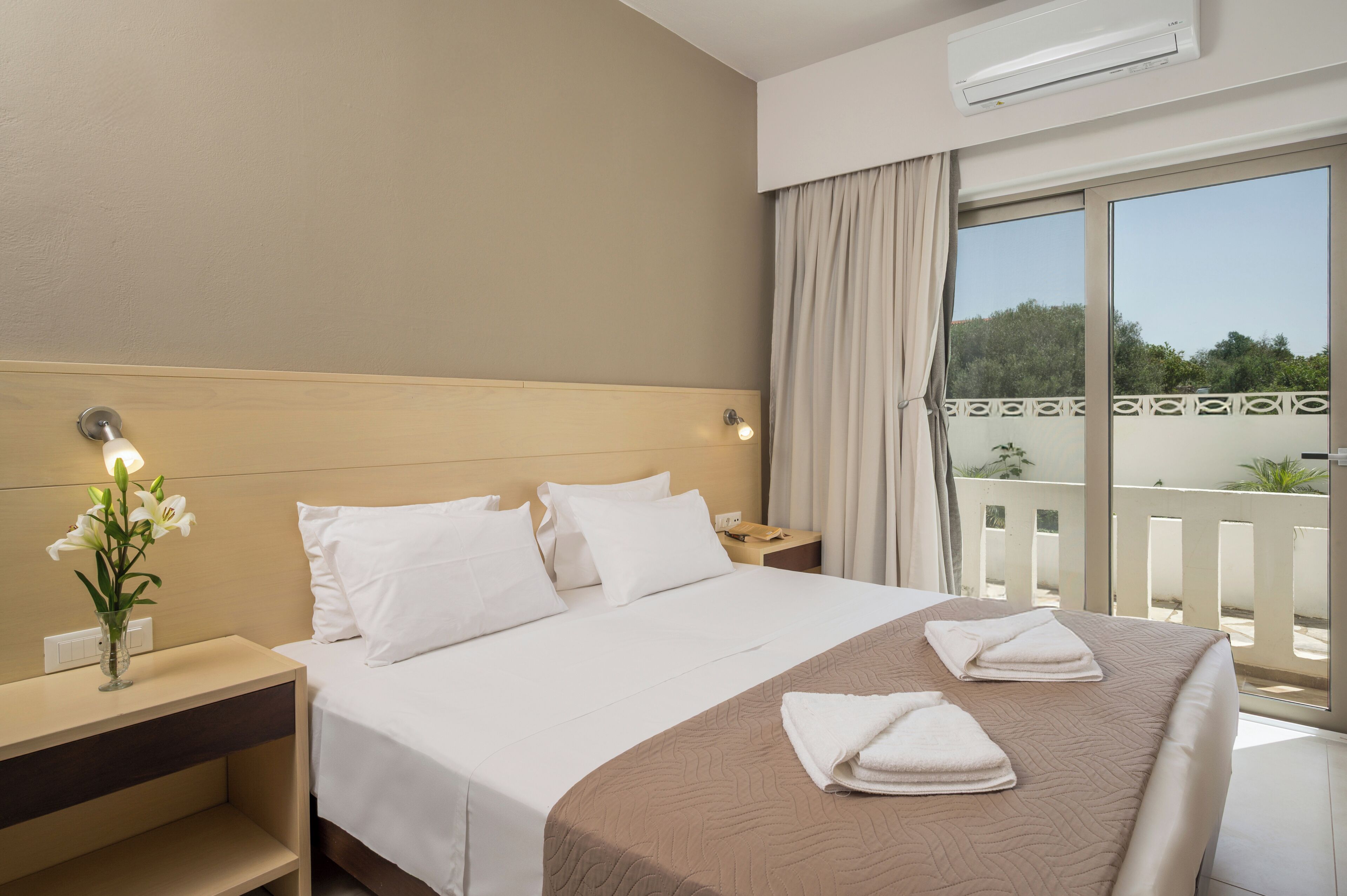 Habitación de hotel con una cama grande, una mesita de noche, un jarrón con flores y vistas al exterior desde un balcón.