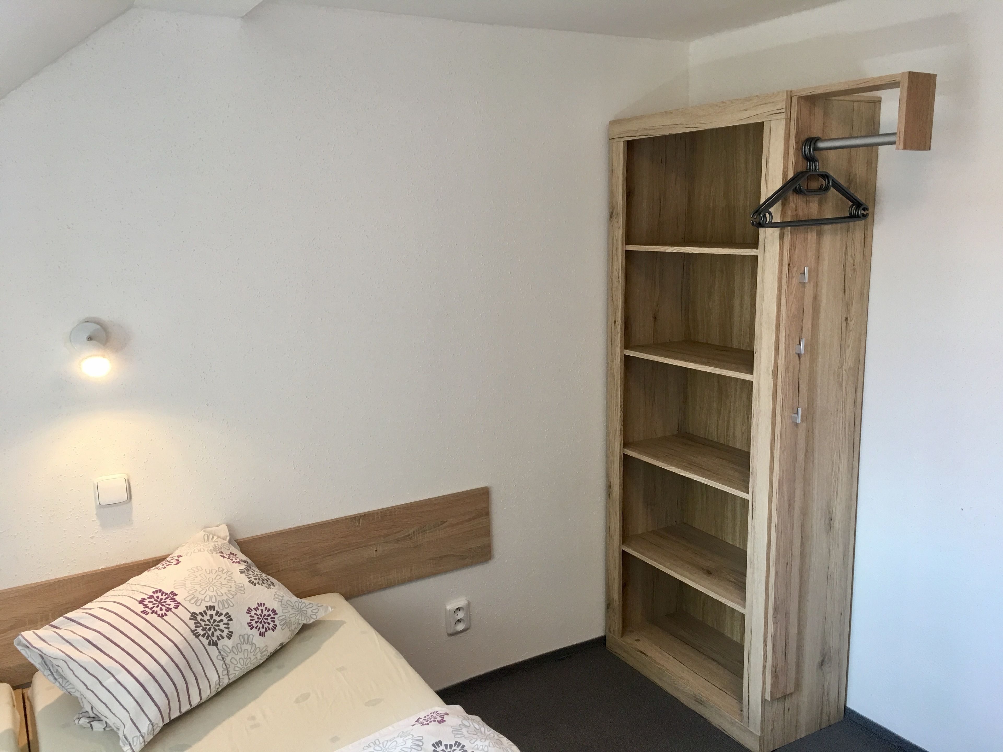 Een slaapkamer met een bed, een houten garderobe met planken, een wandlamp en een stopcontact.