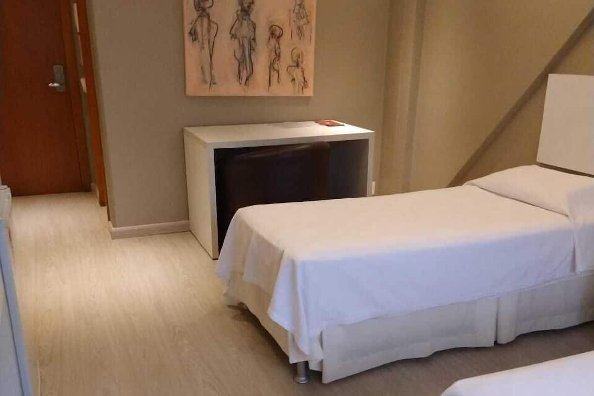 A hotel room with two beds, a desk, a chair, a painting on the wall, and a door.