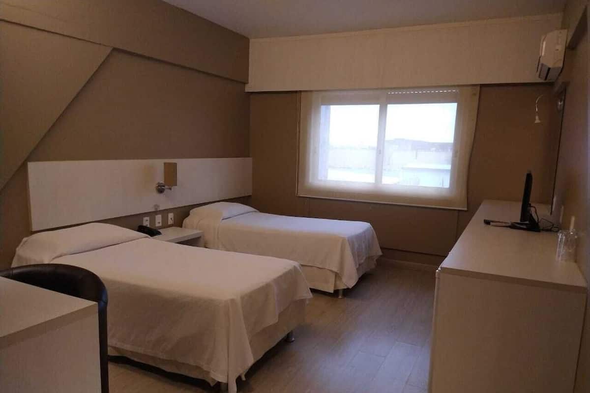 A hotel room with two single beds, a desk, a chair, and a window with a view.