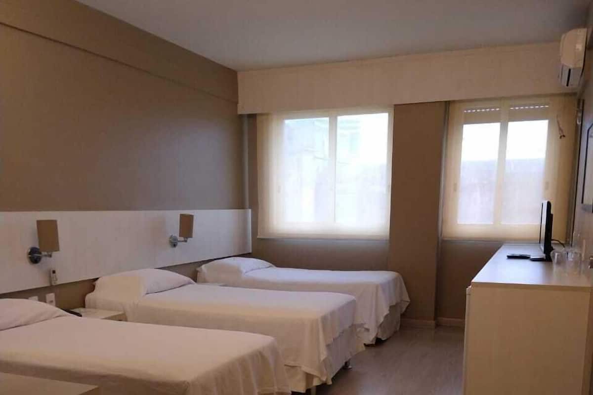A hotel room with three single beds, a desk, and a window with blinds.