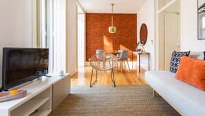 A modern living room with a flat-screen TV, a sofa with an orange cushion, and a dining area with a table and chairs.