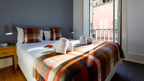 A hotel room with a bed, pillows, a blanket, a nightstand, a lamp, and a view of the outside.