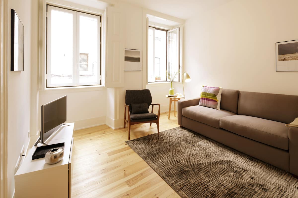 Ein Wohnzimmer mit Sofa, Sessel, Fernseher auf einem Schrank und einem Fenster mit Blick.