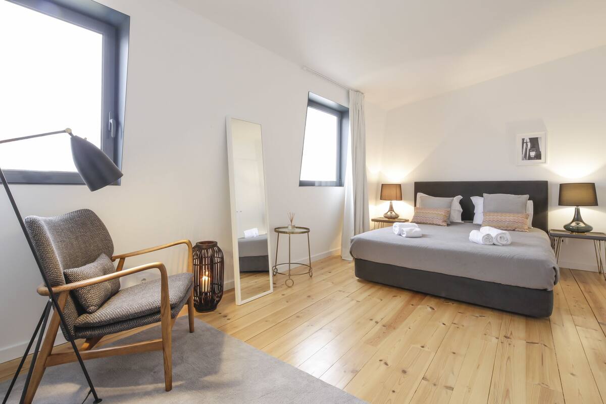 A modern bedroom with a large bed, a chair, a desk lamp, and a mirror.