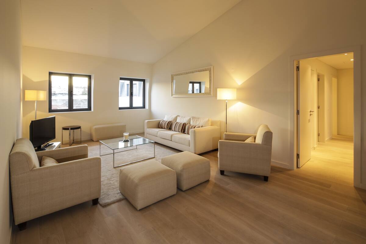 A modern living room with a sofa, armchairs, a coffee table, and a TV. There are windows and a mirror on the walls, and a door leading to another room.