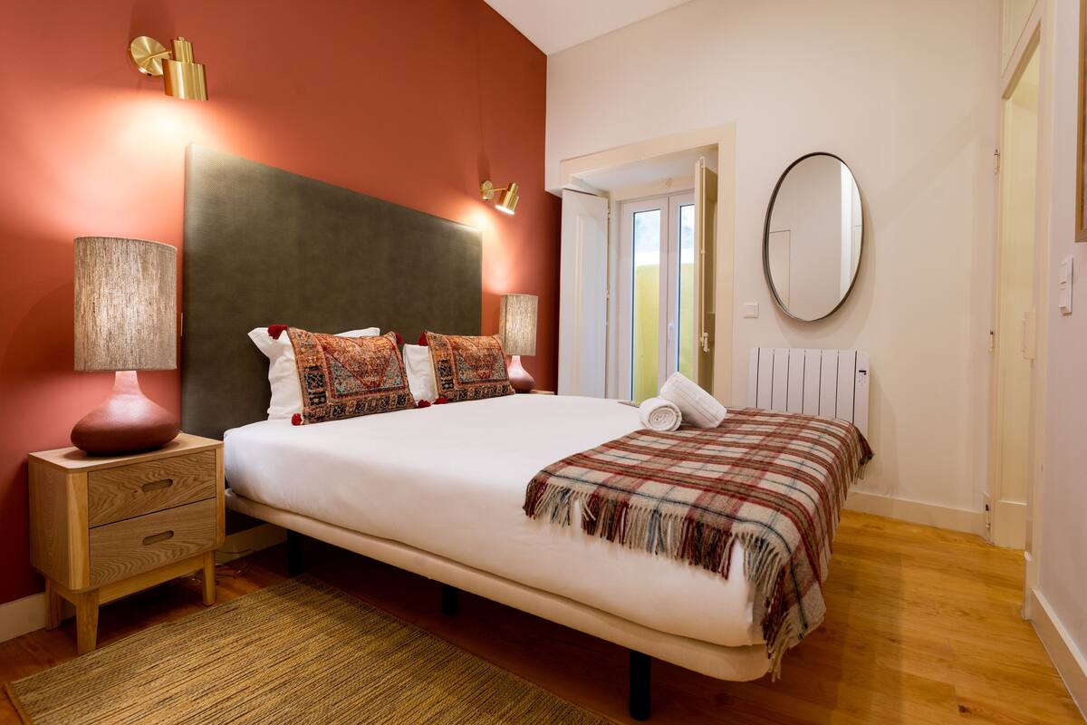 A bedroom with a large bed, bedside lamps, a wooden nightstand, a mirror, and a radiator.