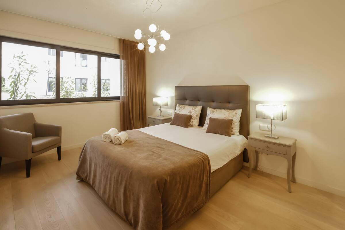 A hotel room with a large bed, a chair, a nightstand, and a window with curtains.