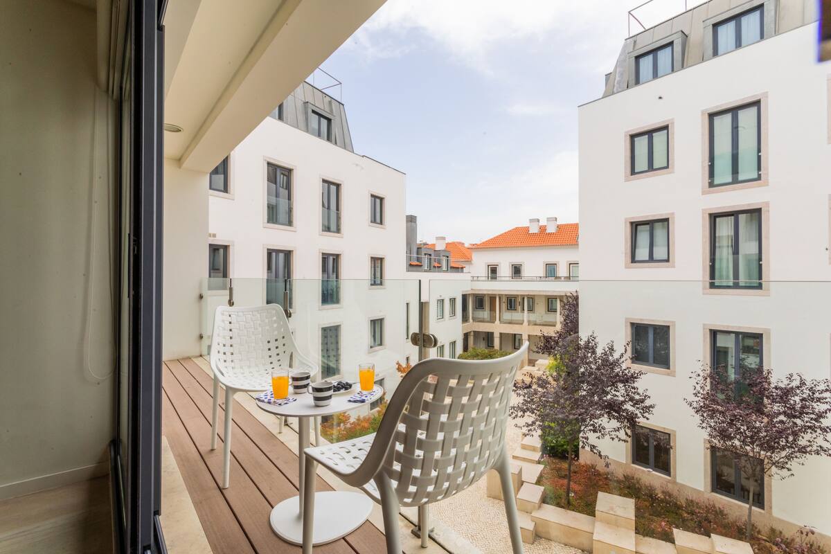A balcony with a table and chairs, overlooking a courtyard with buildings and trees.