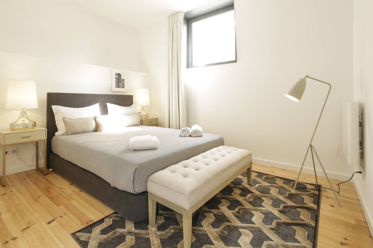 A neatly arranged bedroom with a bed, bedside tables, a bench, a floor lamp, and a window.