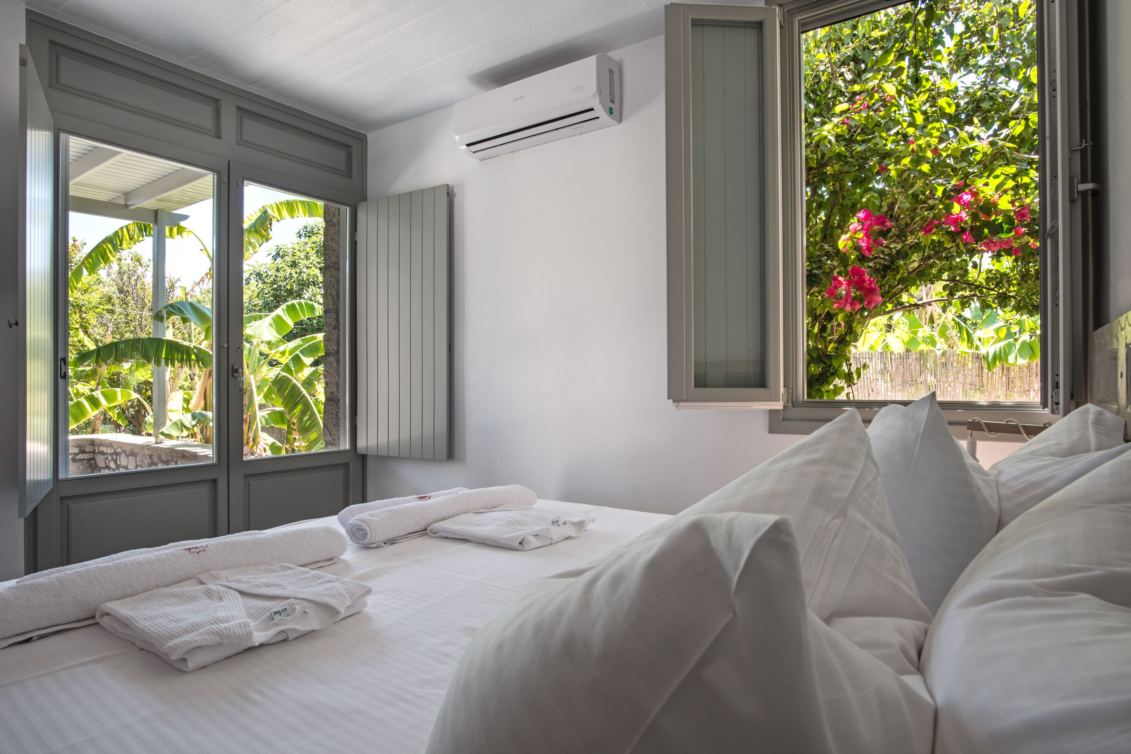 Habitación de hotel con cama, ventana con vistas a follaje y flores, unidad de aire acondicionado y toallero.