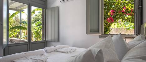 Habitación de hotel con cama, ventana con vistas a follaje y flores, unidad de aire acondicionado y toallero.