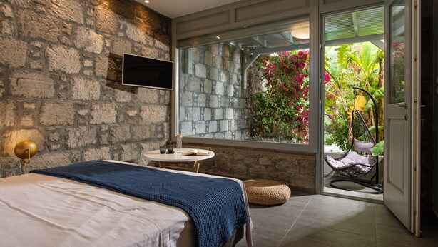 A bedroom with a stone wall, a bed with a blue blanket, a bedside table with a lamp, and a view of a garden through a glass door. - Melagrana (Milos)