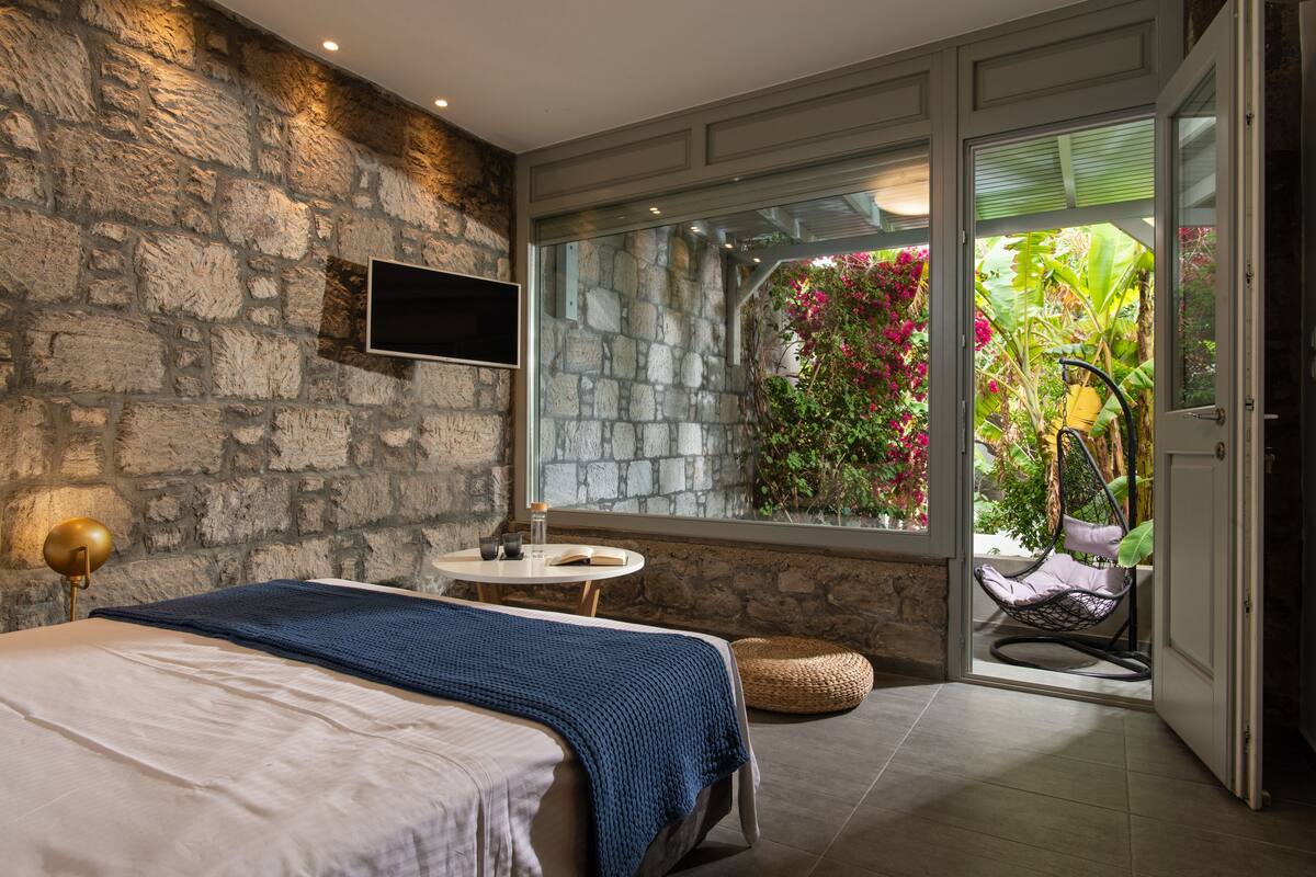 Un dormitorio con pared de piedra, una cama con manta azul, una mesita de noche con lámpara y vista a un jardín a través de una puerta de cristal.