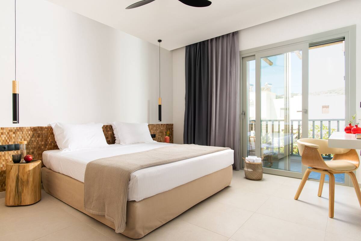 Habitación de hotel moderna con una cama grande, una zona de comedor con mesa y sillas, y un balcón con vistas.