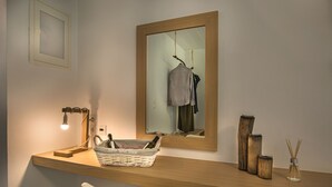 A bathroom with a wooden vanity, a mirror, a hanging robe, a basket with toiletries, and a white chair. - Melagrana (Milos)