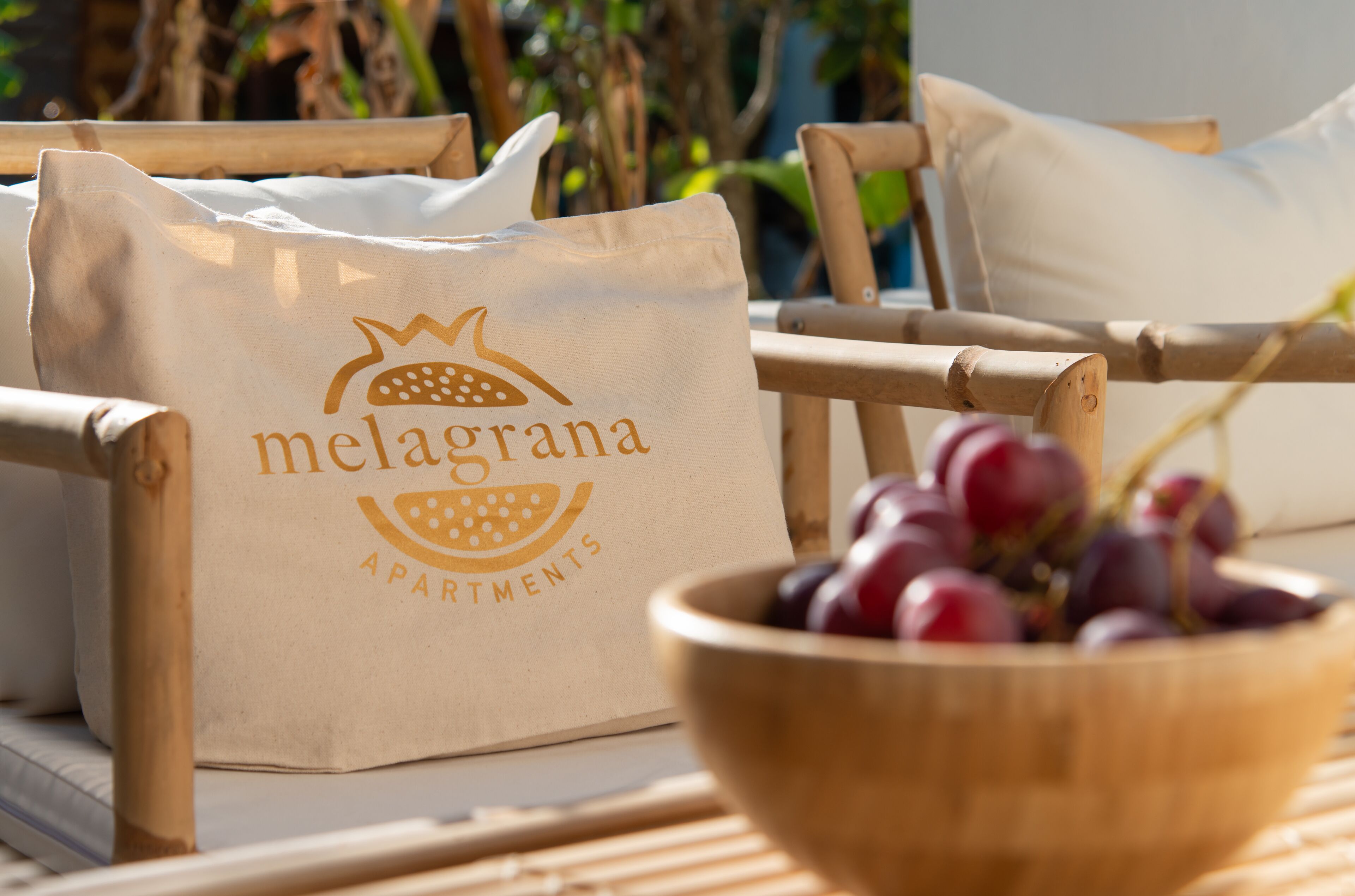 A wooden chair with a white cushion, a wooden bowl with red grapes, and a beige paper bag with the text "melagrana apartments".