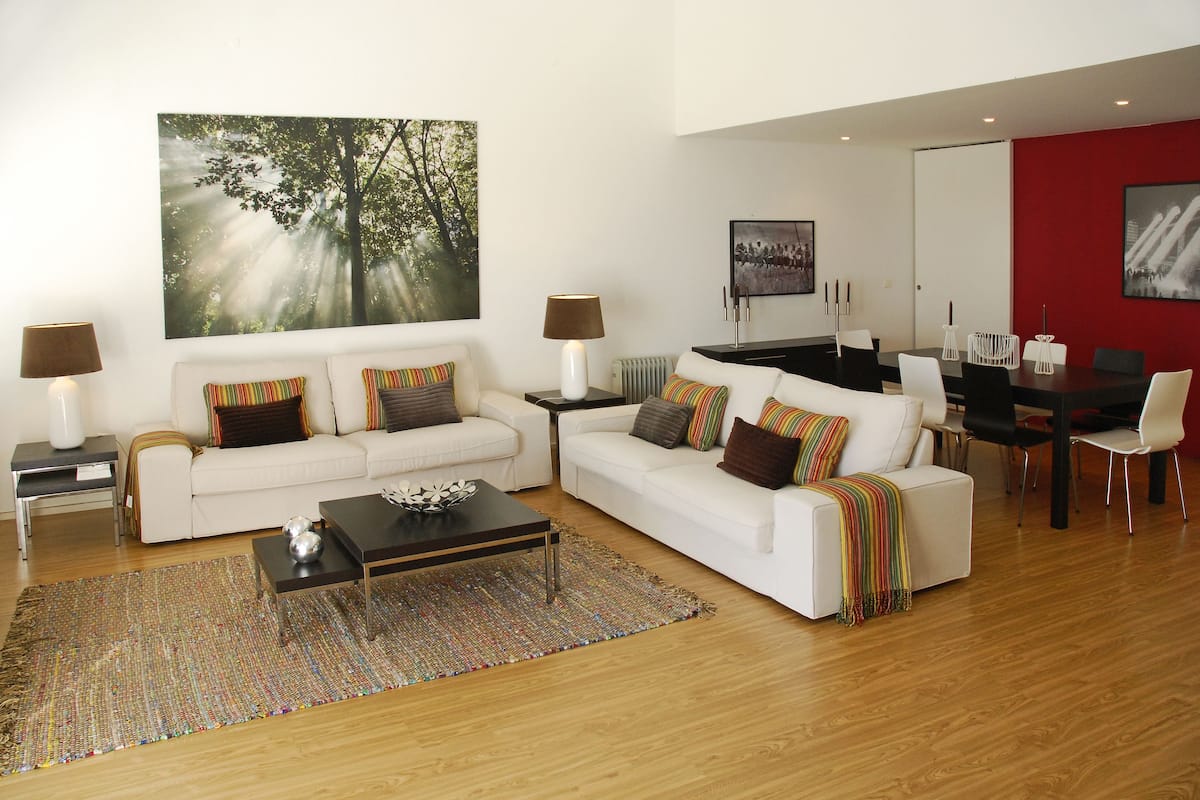 A modern living room with two white sofas, a coffee table, and a dining area with a red accent wall.
