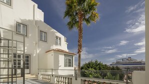 Un bâtiment blanc doté d’un balcon, avec un palmier et un navire de croisière en arrière-plan.