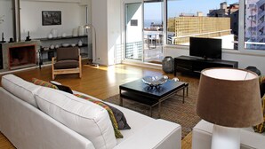 Un salon moderne avec un canapé, un fauteuil, une télévision et une vue sur la ville grâce à de grandes fenêtres.