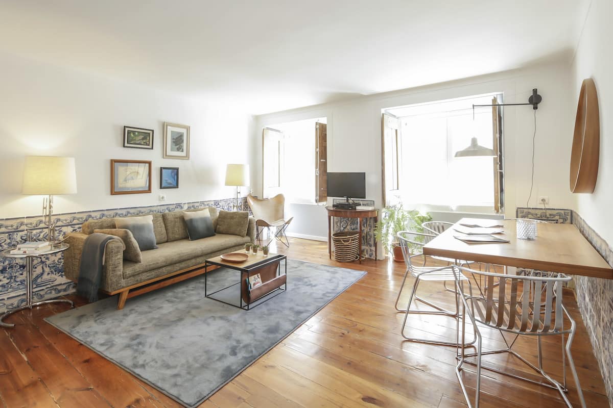 A living room with a sofa, coffee table, and dining area with chairs. There are framed pictures on the wall, a TV, and a lamp.